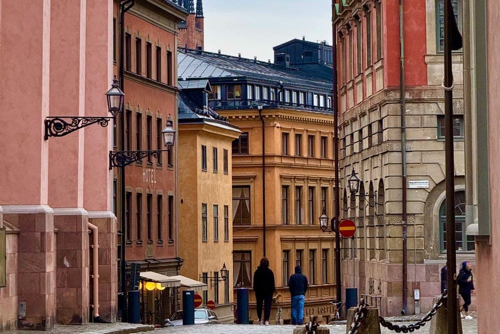 A view down a sloping street in Stockholm, Sweden, with colorful historic buildings on either side and church spires in the distance.