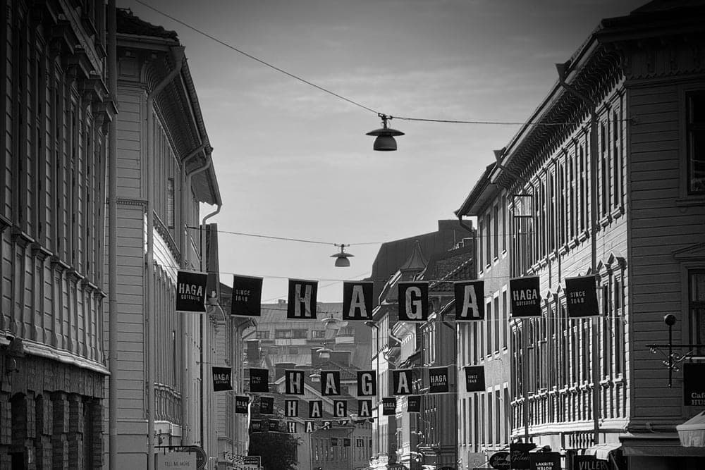 A black and white view of the Haga district in Göteborg, Sweden, with 'HAGA' banners strung across a cobblestone street lined with historic buildings and bustling with pedestrians.