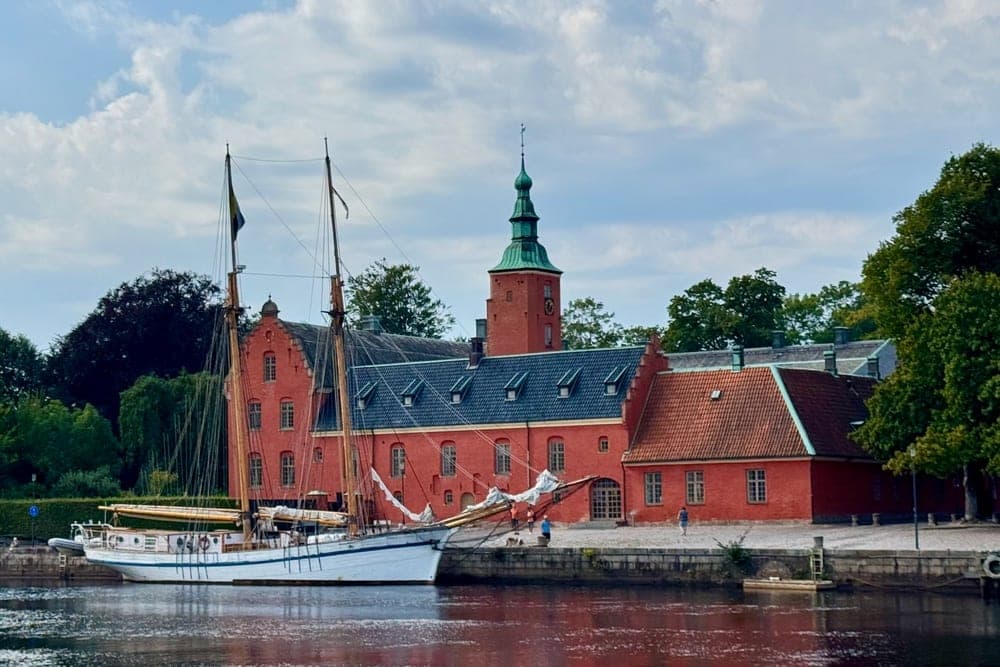A tall ship docked in front of a red historic building with a green tower in Halmstad, Sweden.