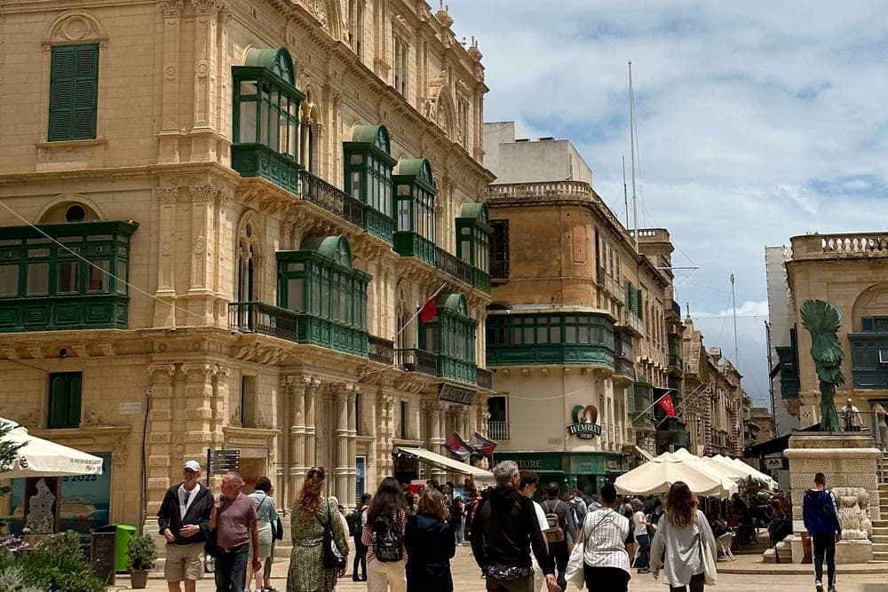 A busy street scene in Valletta, Malta, with people walking past a grand building featuring many traditional green wooden balconies.