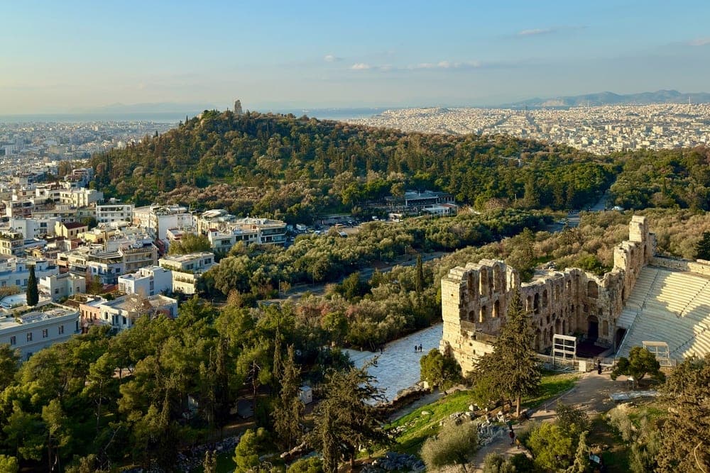 Panoramic view of Athens, Greece, showing ancient ruins in the foreground and the cityscape stretching to the horizon under a clear blue sky.