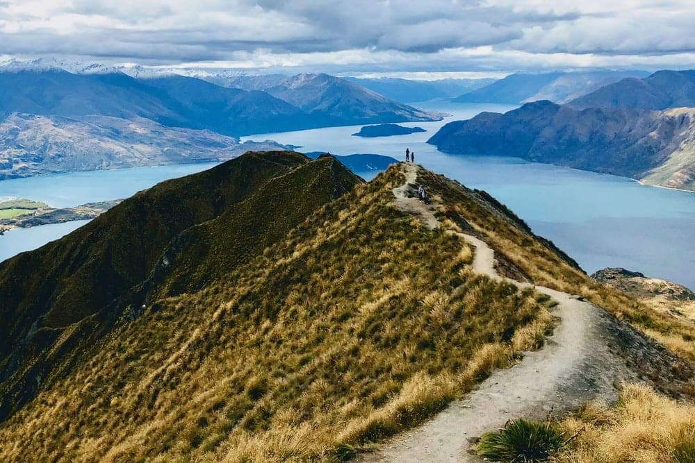 Two hikers stand on a narrow ridge path at the summit of Roys Peak, overlooking Lake Wānaka and surrounding mountains in New Zealand.