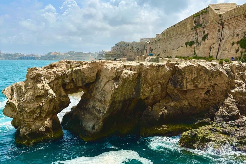 Natural rock arch in the Mediterranean Sea near Valletta, Malta, with ancient fortifications in the background under a blue sky with clouds.
