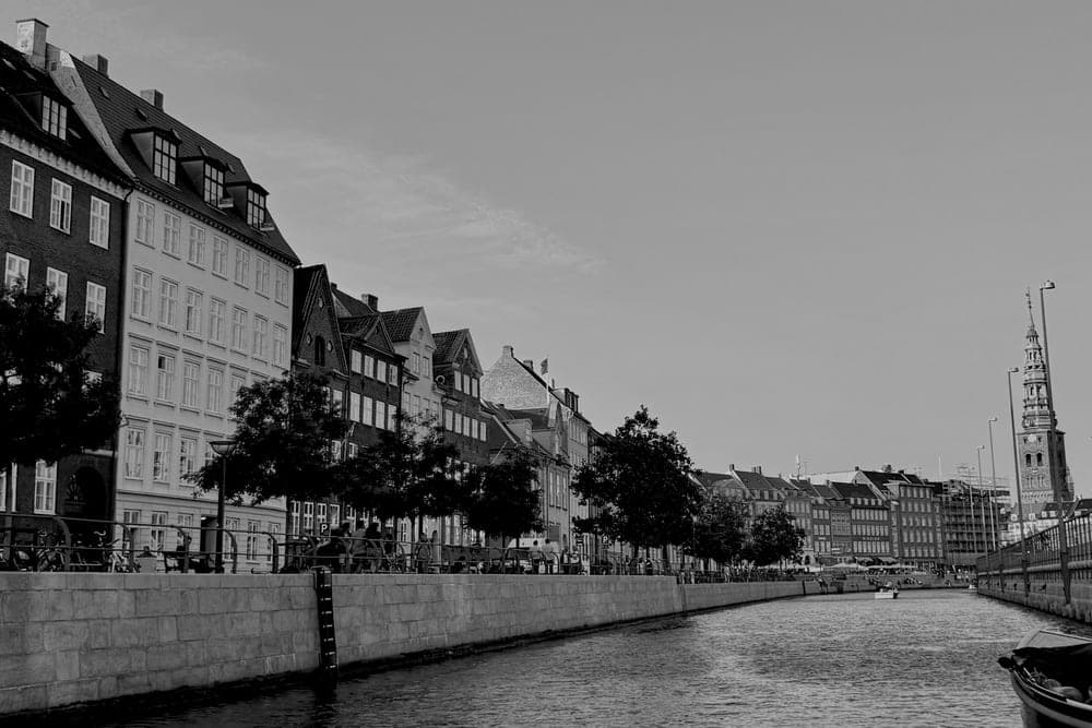 A wide view of the Nyhavn canal in Copenhagen, Denmark, with historic buildings lining the waterfront and a distant church spire.