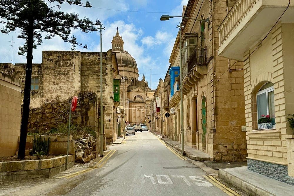 A street in Xewkija, Malta, leading towards the dome of the Basilica of Saint John the Baptist, with traditional stone buildings and a large tree.