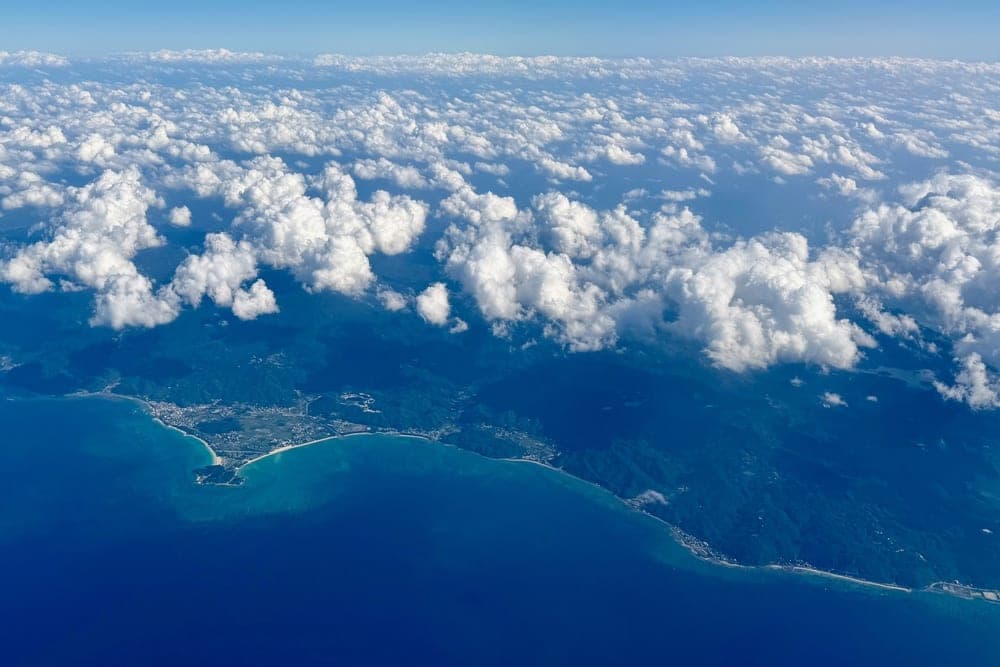 Aerial view of the coast of Okinawa, Japan, showing the deep blue ocean meeting a green, mountainous coastline dotted with villages, under a sky filled with white clouds.