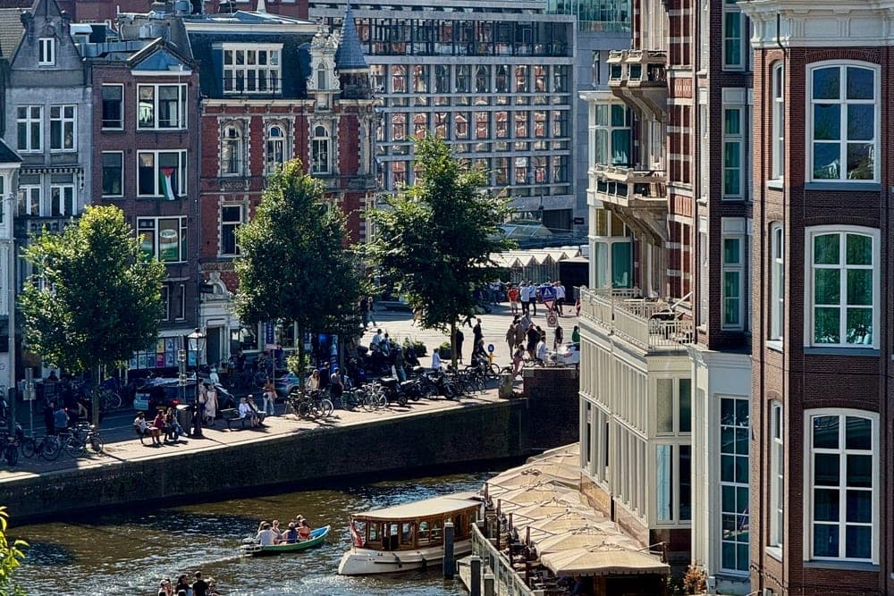 A sunny afternoon view of a canal in Amsterdam, Netherlands, with boats, historic buildings, and pedestrians.