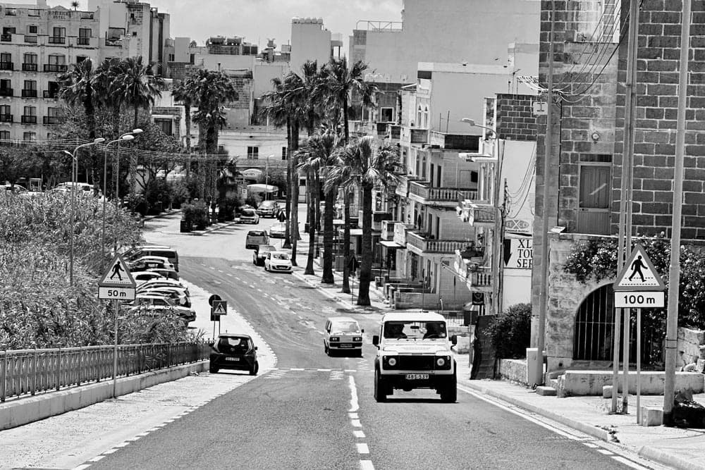 A black and white image of a road in Xlendi, Munxar, Malta, with palm trees, cars, and buildings.