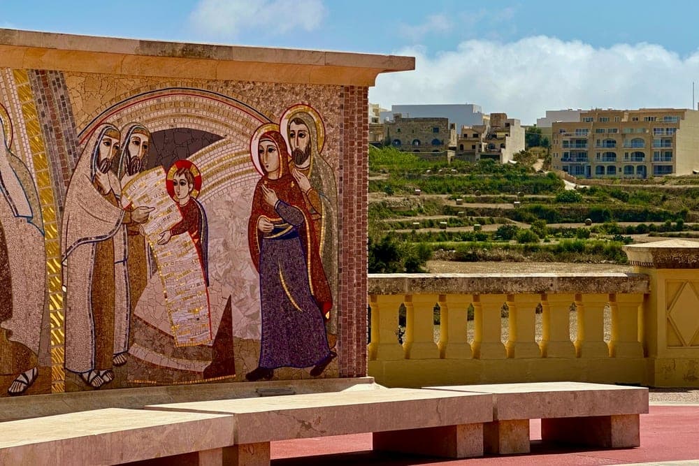 A colourful mosaic depicting religious figures on a wall in Għarb, Malta, with terraced hills and buildings in the background under a bright blue sky.