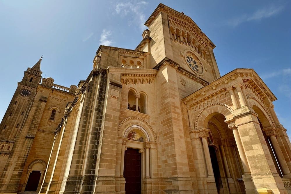 The Ta' Pinu Basilica in Għarb, Malta, seen from a low angle against a bright blue sky.