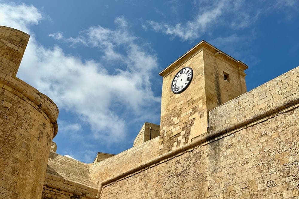 The clock tower of Gharsi, Malta, stands tall against a blue sky with white clouds, adjacent to ancient stone fortifications.