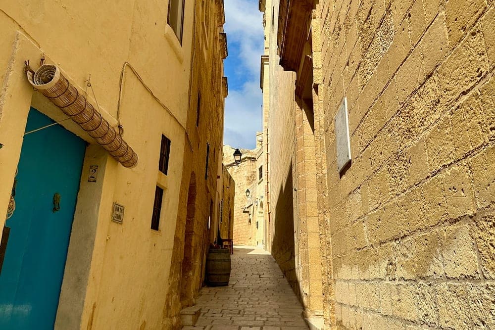A narrow cobblestone street in Victoria, Gozo, Malta, flanked by tall, sunlit limestone buildings, leading to a distant church.