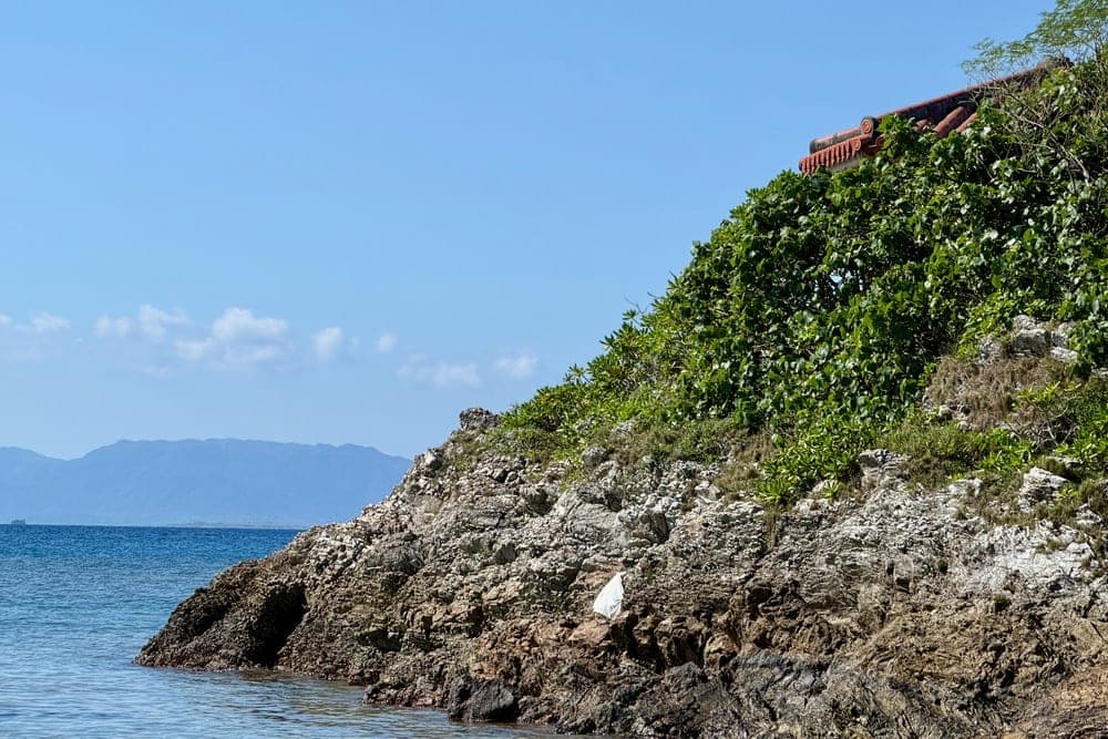 A rocky coastline in Taketomi, Japan, with lush green foliage and a traditional tiled roof under a clear blue sky and calm sea.