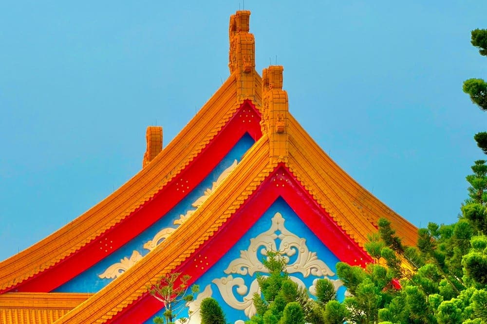 Close-up of the ornate golden-yellow and red roofs with blue and white decorative panels of the National Chiang Kai-shek Memorial Hall in Taipei, Taiwan, framed by green trees.