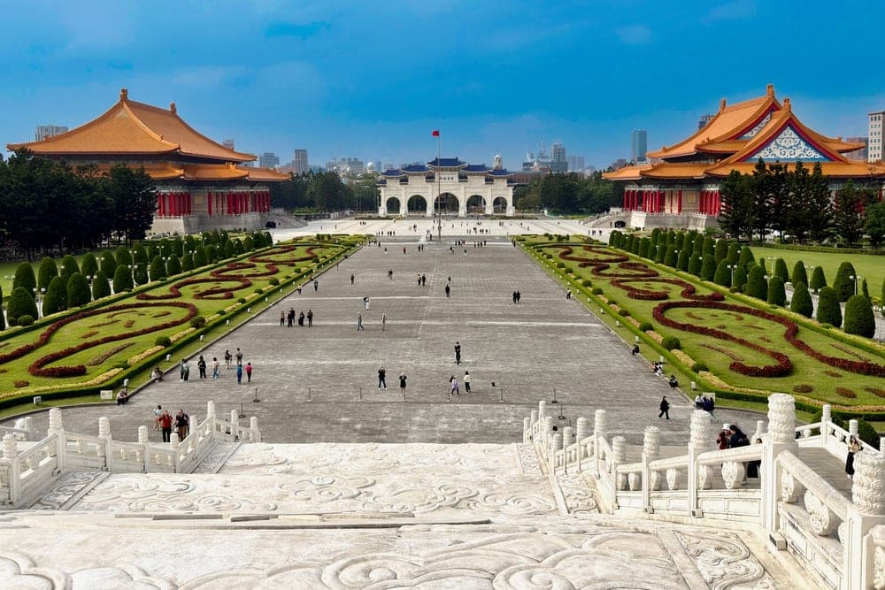 The National Chiang Kai-shek Memorial Hall in Taipei, Taiwan, viewed from ornate stone steps, with a large plaza, flanking buildings, gardens, and a distant cityscape under a blue sky.