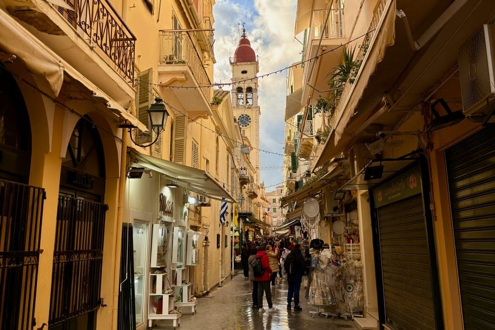 A street in Corfu, Greece, with yellow buildings, balconies, shops, and a prominent bell tower with a red dome.