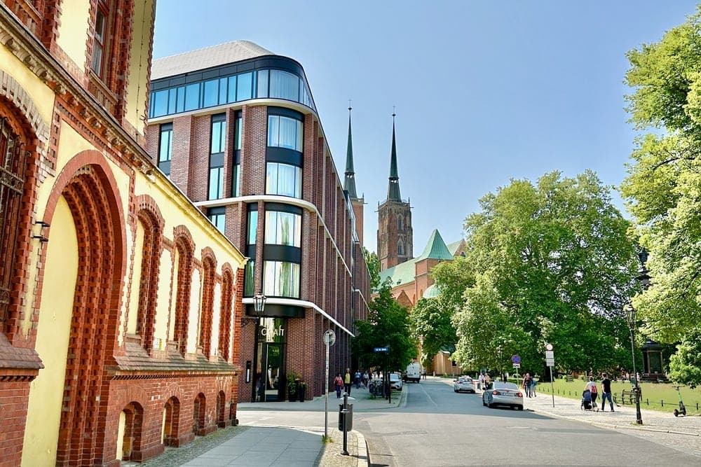 A street scene in Wrocław, Poland, showing historic brick buildings, modern architecture, and Gothic cathedral spires under a clear blue sky.