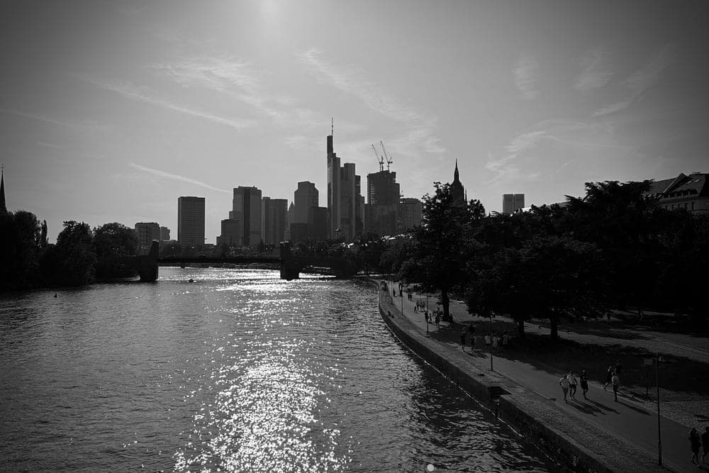 Frankfurt skyline at dusk across the Main River, Germany, with sunlit reflections on the water and people walking along the promenade.