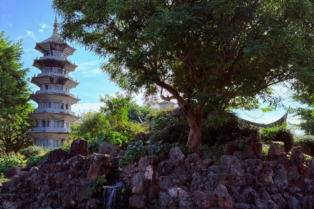 A stone pagoda stands in a lush garden with a waterfall and pond in Okinawa, Japan.