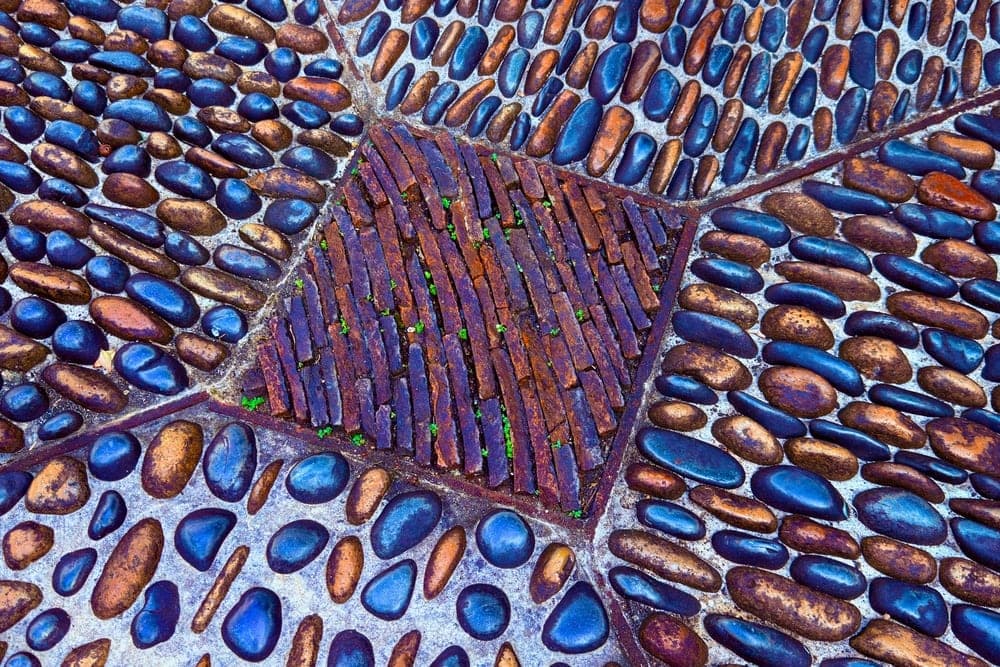 Close-up of a mosaic pathway in Okinawa, Japan, with smooth blue and orange stones surrounding a central diamond of reddish-brown bricks with green grass.