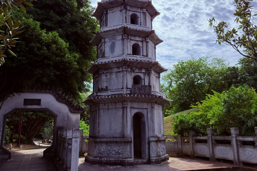 A tall, grey stone pagoda with multiple tiers and ornate roofs, set amidst lush green trees in Okinawa, Japan.