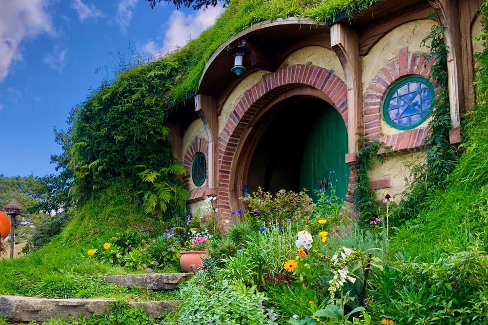 A hobbit hole with a green door and round windows, nestled into a grassy hill in Matamata, New Zealand, surrounded by flowers and stone steps.