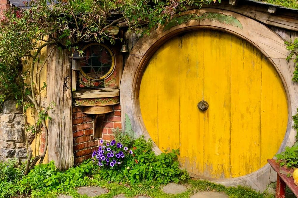 A hobbit-hole with a bright yellow round door in Matamata, New Zealand, surrounded by green grass and flowers.
