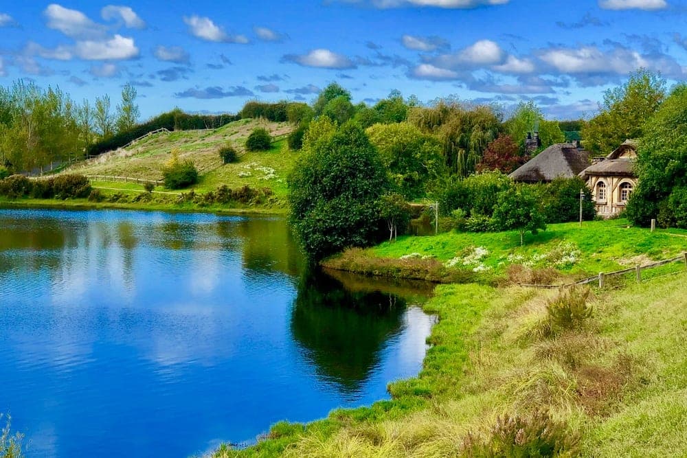 A hobbit-hole dwelling with a thatched roof sits beside a bright blue lake in Matamata, New Zealand.