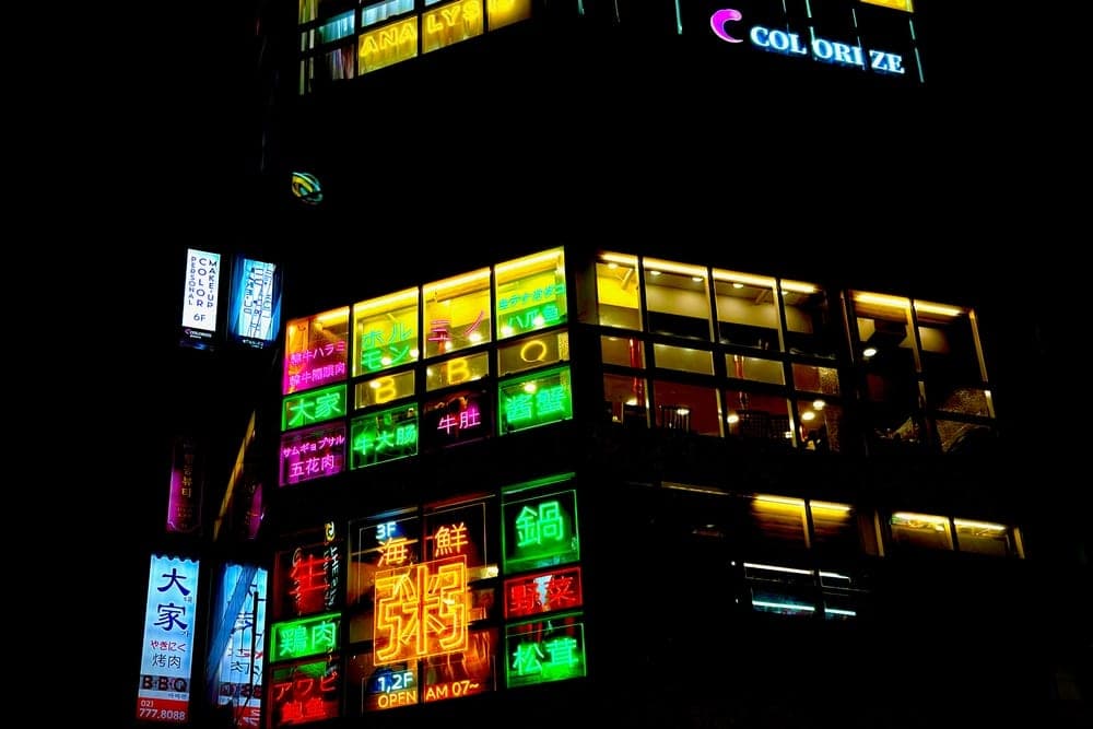 A nighttime view of commercial buildings in Seoul, South Korea, covered in bright, colorful neon signs advertising various businesses like beauty salons, restaurants, and shops.
