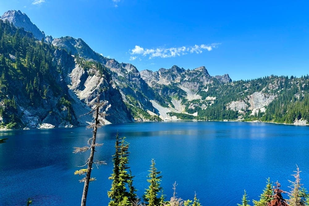 A deep blue alpine lake surrounded by majestic, tree-covered mountains under a clear blue sky in Snoqualmie Pass, Washington, United States.
