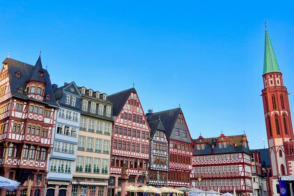 A row of colourful, historic half-timbered buildings in Frankfurt, Germany, with St. Nicholas Church tower to the right, under a clear blue sky.
