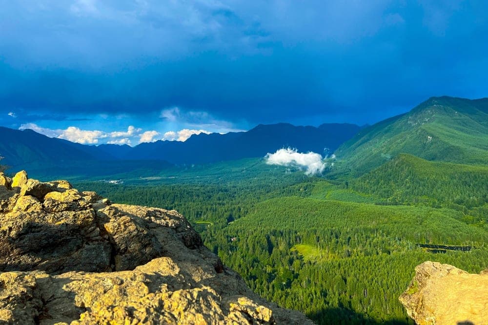 Panoramic view from Rattlesnake Mountain Trail in North Bend, Washington, United States, looking over forested valleys and distant mountains under a dramatic sky.