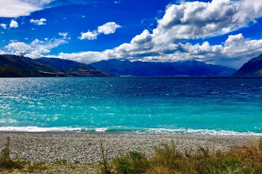 The vibrant turquoise waters of Lake Hāwea, New Zealand, with a pebble beach in the foreground and rugged mountains under a dramatic blue sky with white clouds.