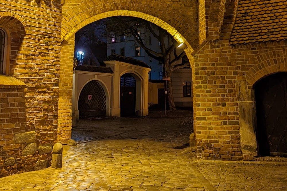 A historic arched brick passageway in Wrocław, Poland, illuminated by warm yellow lights at night, leading to a cobblestone courtyard.