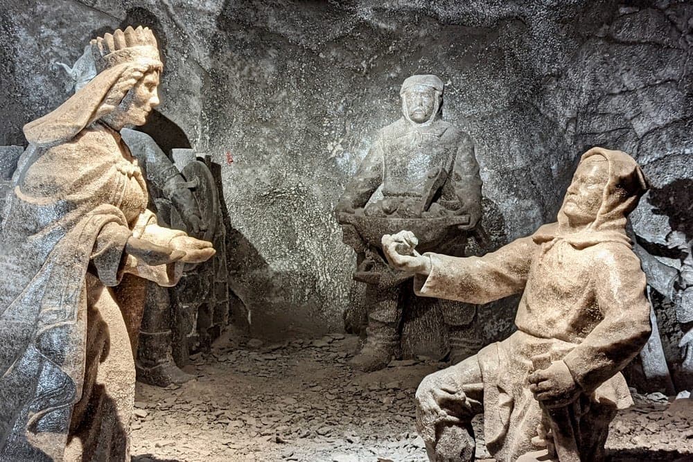 Three salt statues depicting historical figures in a salt mine chamber in Krakow, Poland. A seated miner gestures towards a queen.