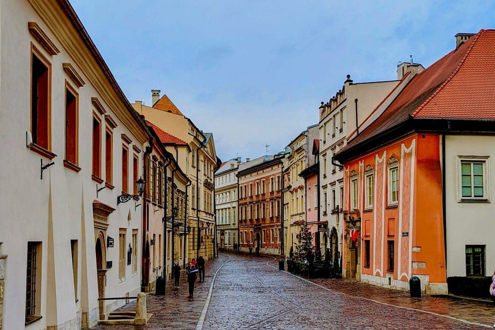 A cobblestone street in Krakow, Poland, lined with historic buildings under a cloudy sky.