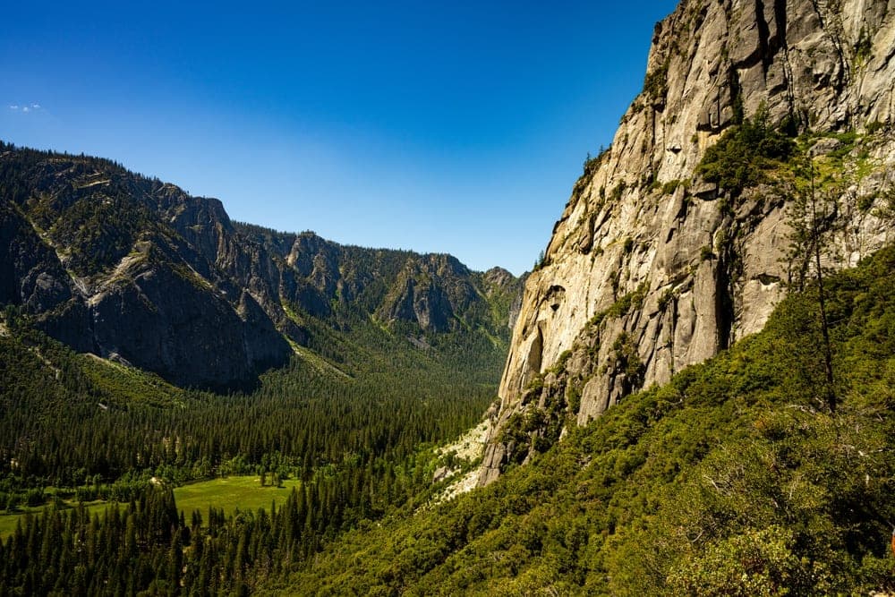 A vast view of Yosemite National Park, California, United States, featuring a towering granite cliff on the right and a deep, forested valley stretching into the distance under a clear blue sky.