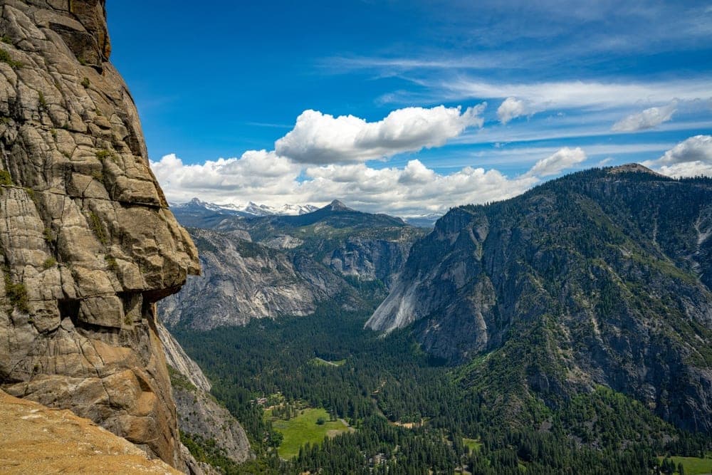 A dramatic view of Yosemite Valley from a high vantage point, with a large granite cliff in the foreground and a vast, tree-filled valley and distant mountains under a blue, cloudy sky.