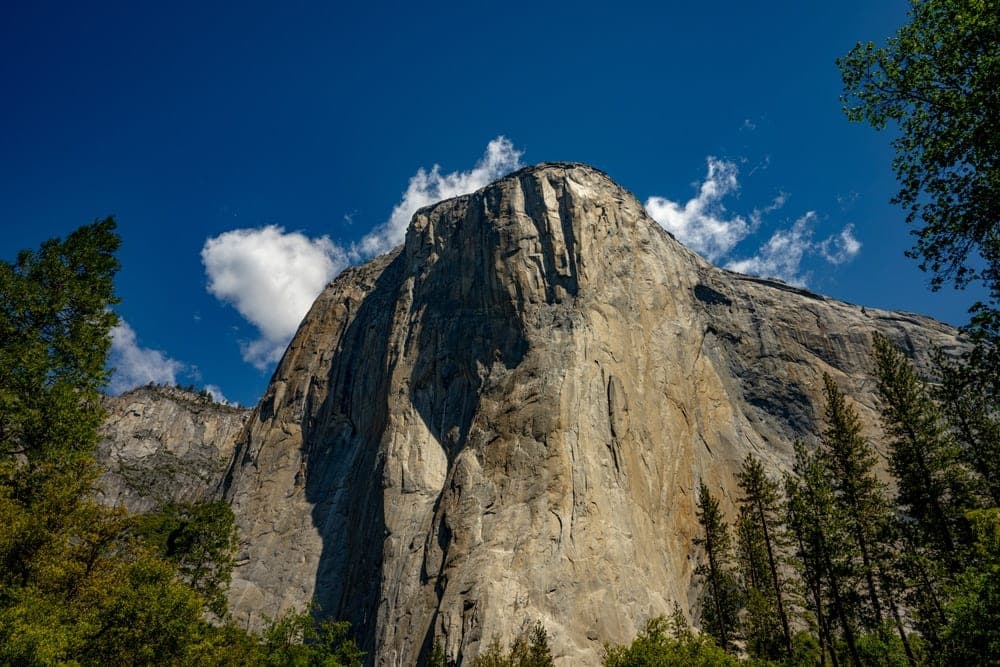 The colossal granite face of El Capitan in Yosemite National Park, USA, seen from below under a bright blue sky with scattered clouds.
