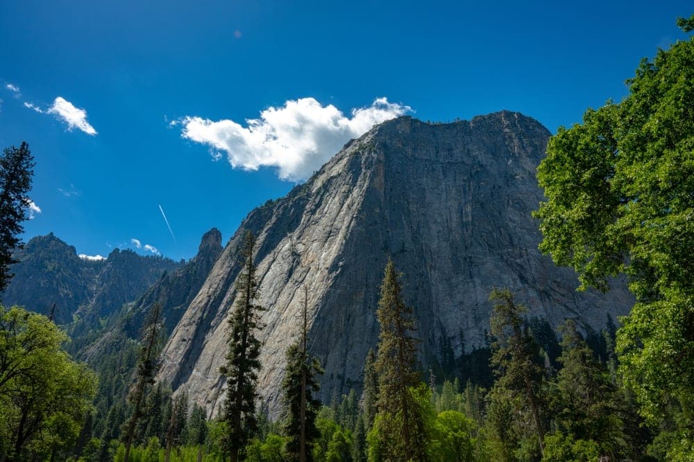 The towering granite face of El Capitan in Yosemite National Park, California, United States, under a bright blue sky with scattered clouds and lush green trees in the foreground.
