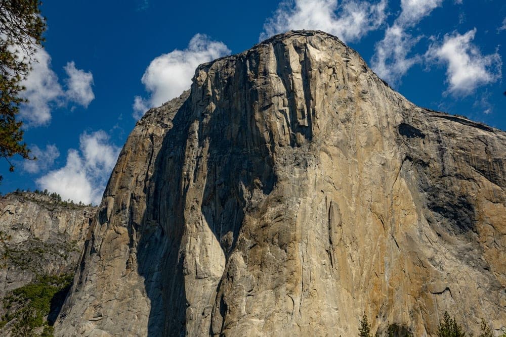 The massive granite face of El Capitan in Yosemite National Park, California, United States, under a bright blue sky with scattered clouds.