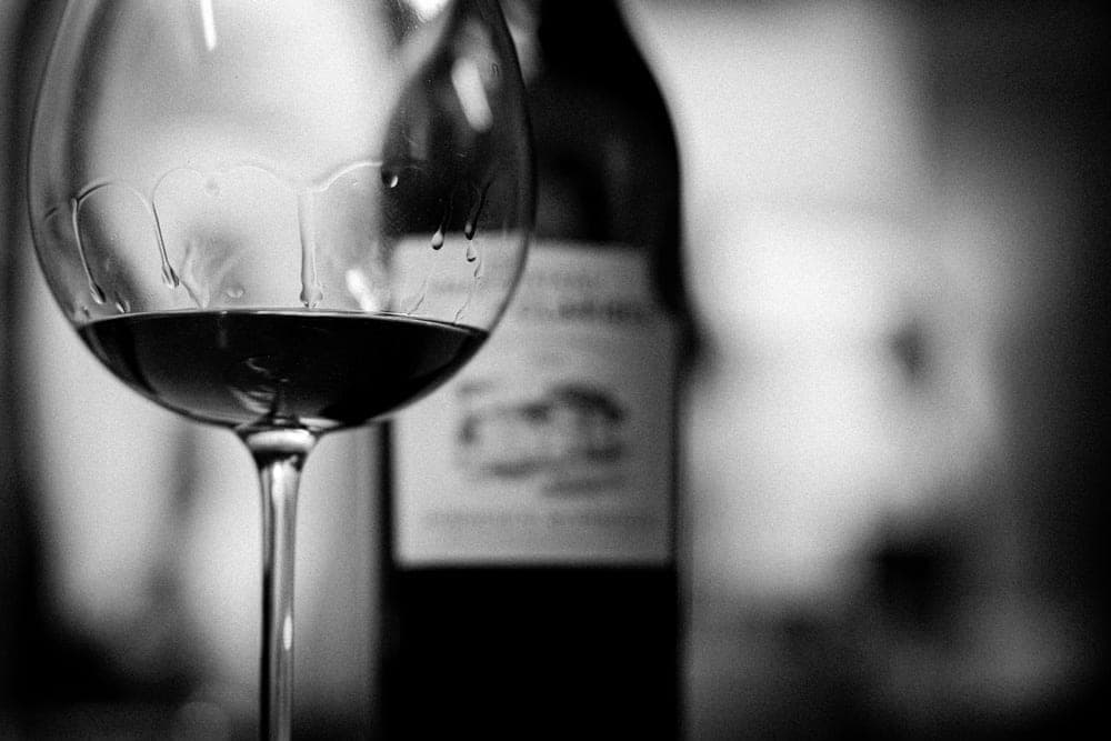 A black and white photograph showing a glass of red wine in sharp focus in the foreground, with a blurred wine bottle in the background. San Francisco, United States.
