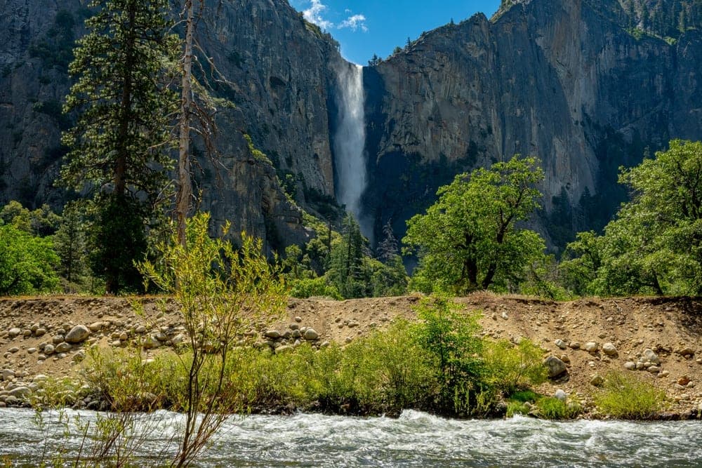 Yosemite National Park, United States, with a tall waterfall plunging down a granite cliff face under a bright blue sky with white clouds, a river in the foreground.