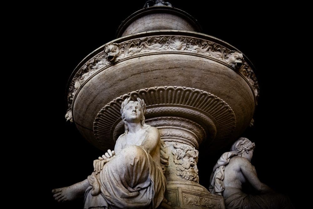 A stone fountain sculpture featuring classical female figures and carved faces in Budapest, Hungary, dramatically lit against a black background.