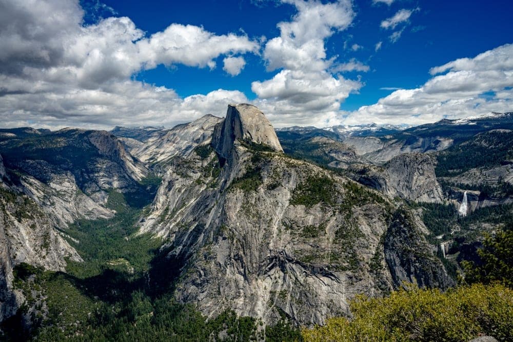 A wide-angle view of Half Dome in Yosemite National Park, California, United States, with Yosemite Falls visible in the distance under a blue sky with white clouds.