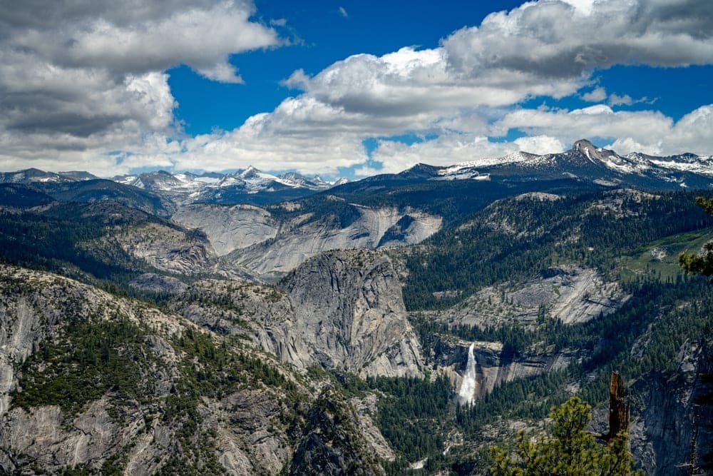 Panoramic view of Yosemite National Park, California, United States, showing granite cliffs, forests, and a waterfall under a blue sky with clouds.