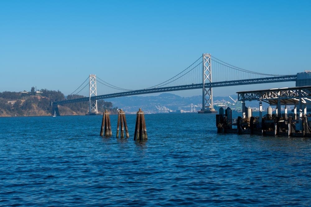 The Bay Bridge spanning the San Francisco Bay in San Francisco, California, United States, under a clear blue sky, with wooden pilings in the foreground and a pier on the right.