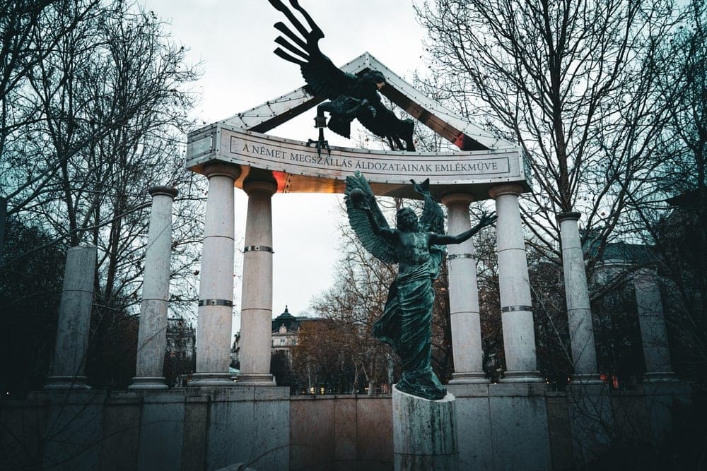 The Monument to the Victims of the German Occupation in Budapest, Hungary, featuring bronze angel statues and winged figures under an overcast sky.