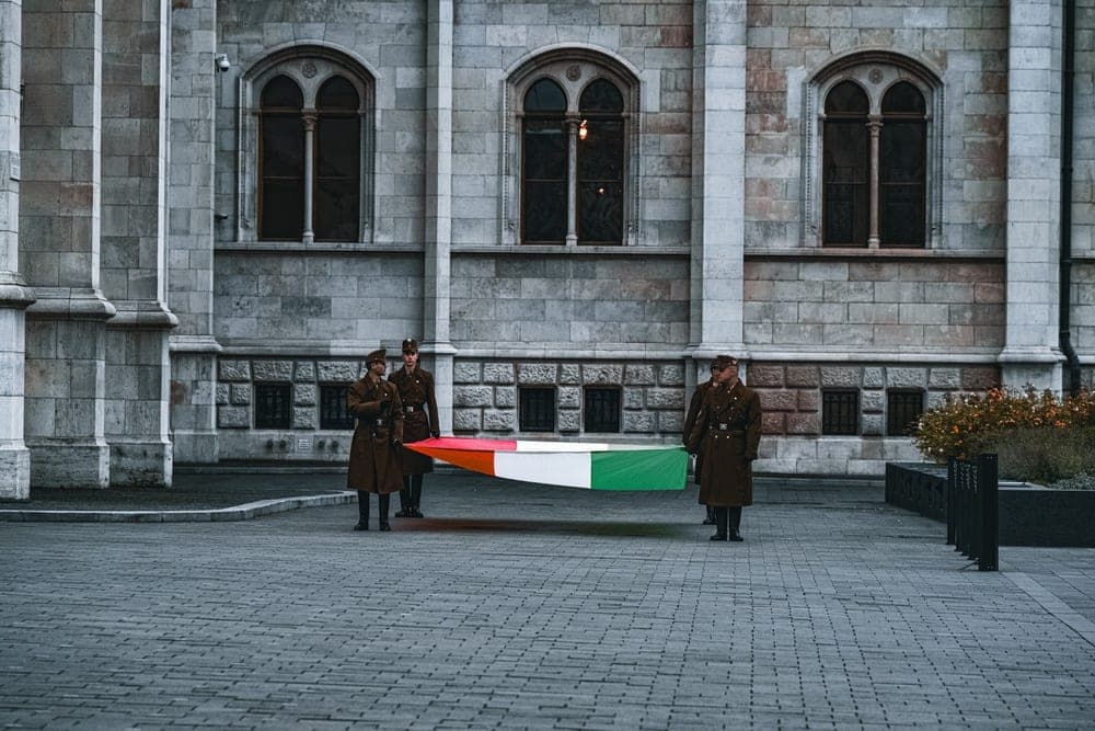 Four guards in uniform carrying the Hungarian flag in front of a grand stone building in Budapest, Hungary.