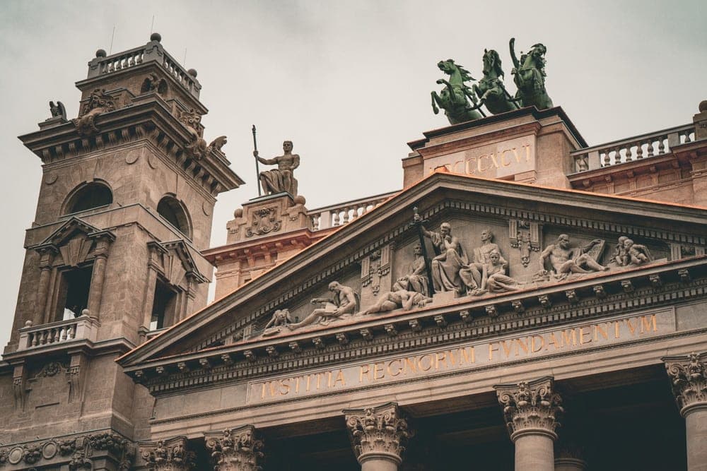 Ornate classical architecture of a building in Budapest, Hungary, featuring statues, reliefs, and inscriptions, under an overcast sky.